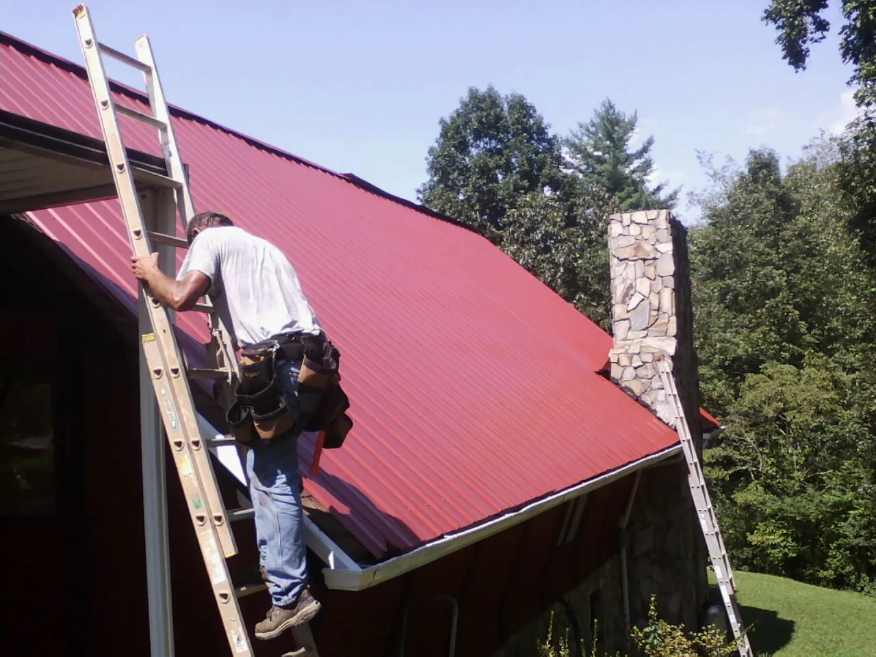 Roofer on a ladder beside a red metal roof and stone chimney — Soffit & Fascia Repair by Roofing Brothers in Henefer